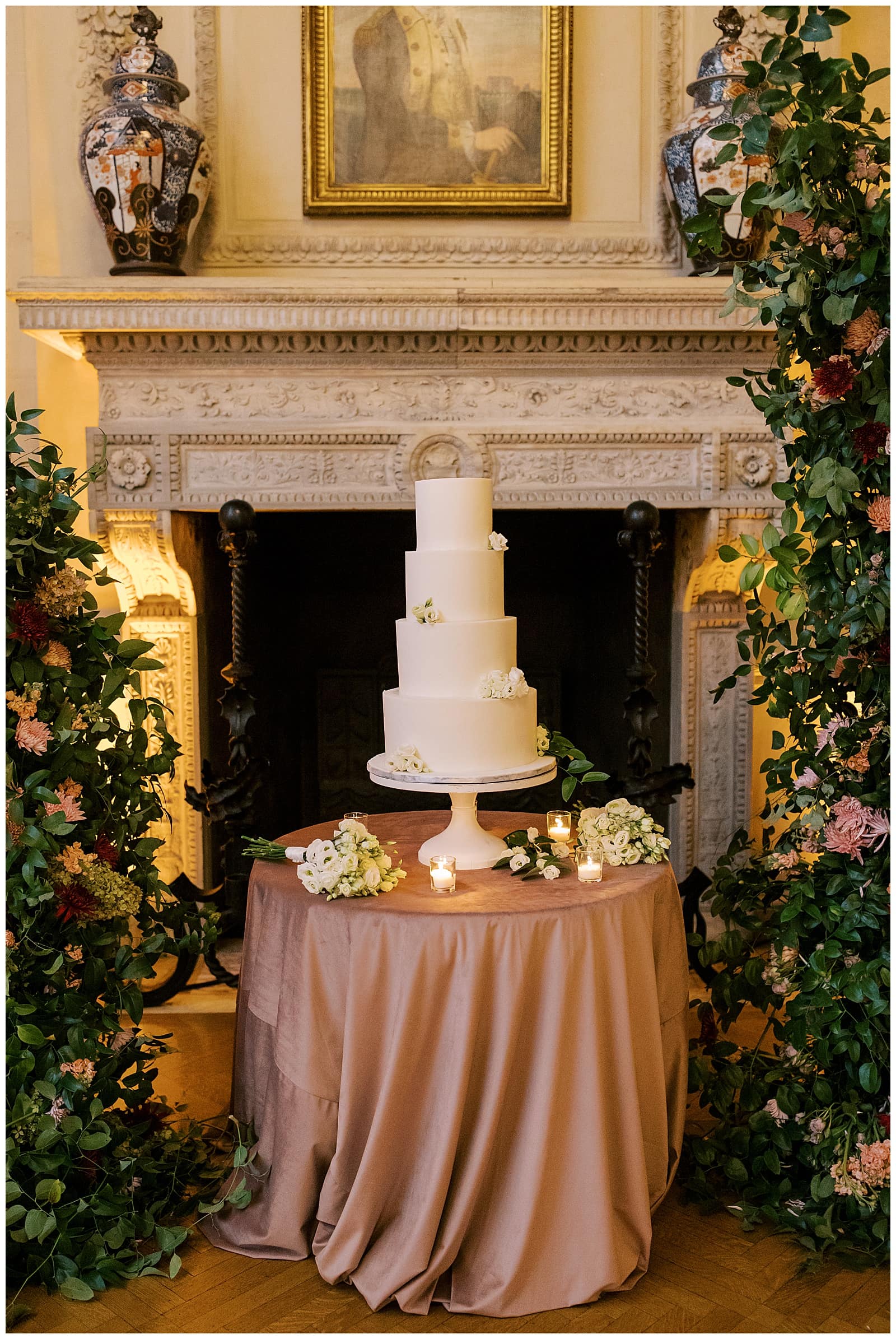 Image of a four tier white wedding cake on a table with velvet tablecloth in between two large floral arrangements in front of the fire place at the Anderson House