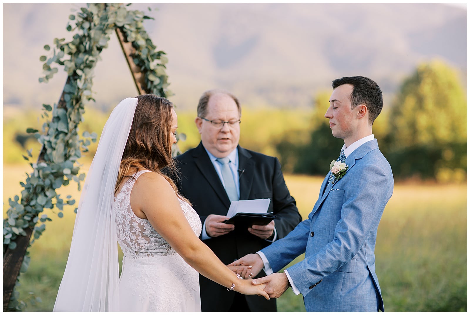 A groom in a periwinkle colored tux and bowtie stands across from a bride in a white dress as they hold hands with a priest behind them reading from a book in the middle of a field with the Smoky mountains in the backdrop