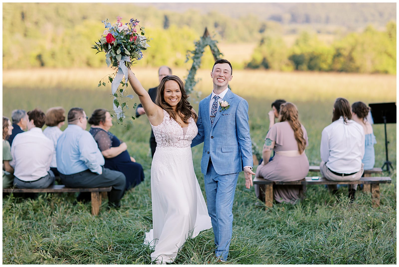 A woman in a wedding dress holds a bouquet high above her head while holding hands with a groom in a tux smiling straight at the camera in the middle of a field in the smoky mountains