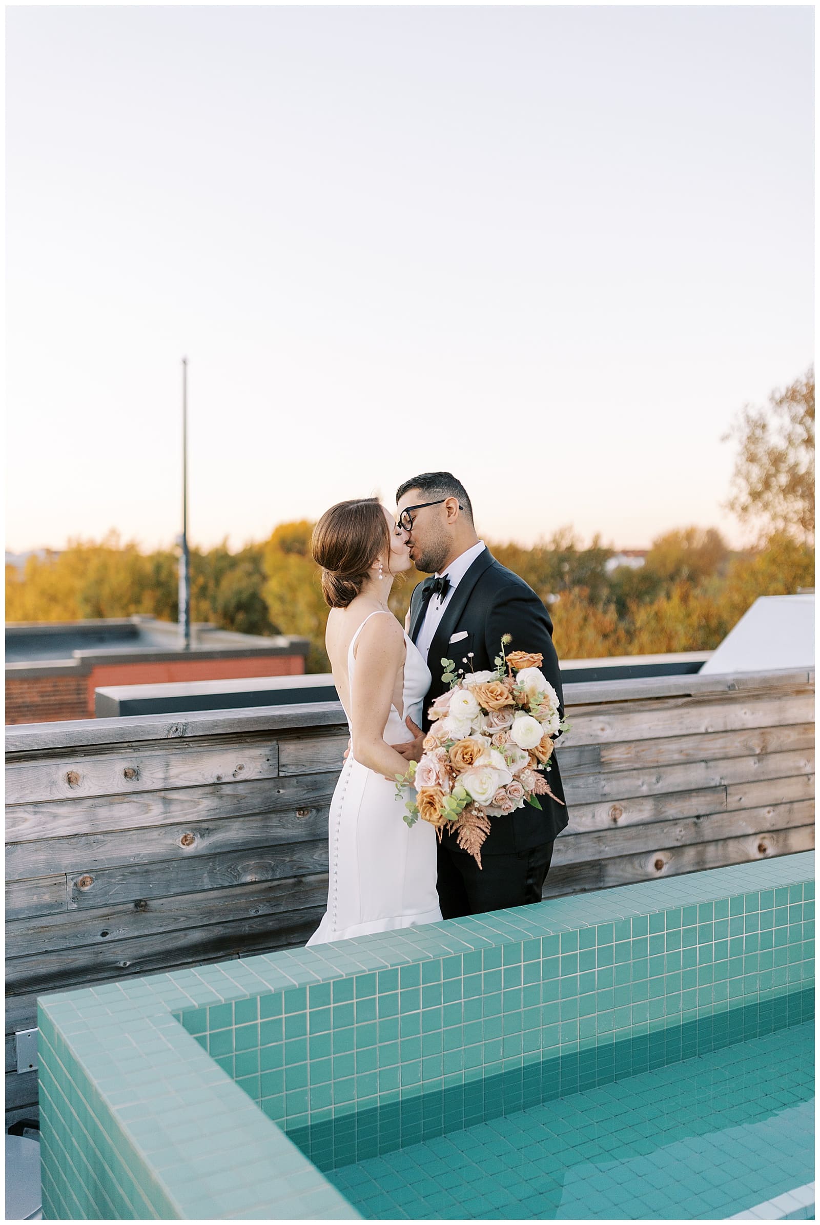 Young redheaded bride in a wedding gown kissing a handsome groom in a black tux by a turquoise colored pool on the rooftop of the Common House Richmond