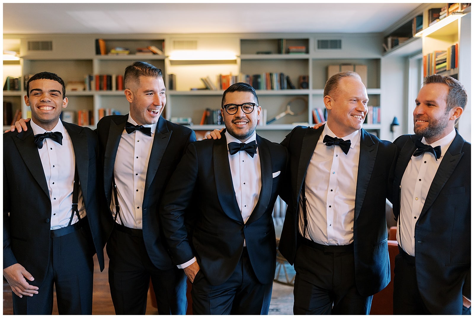 Five men in black tuxes with black bow ties laugh together in the library of the Common House Richmond