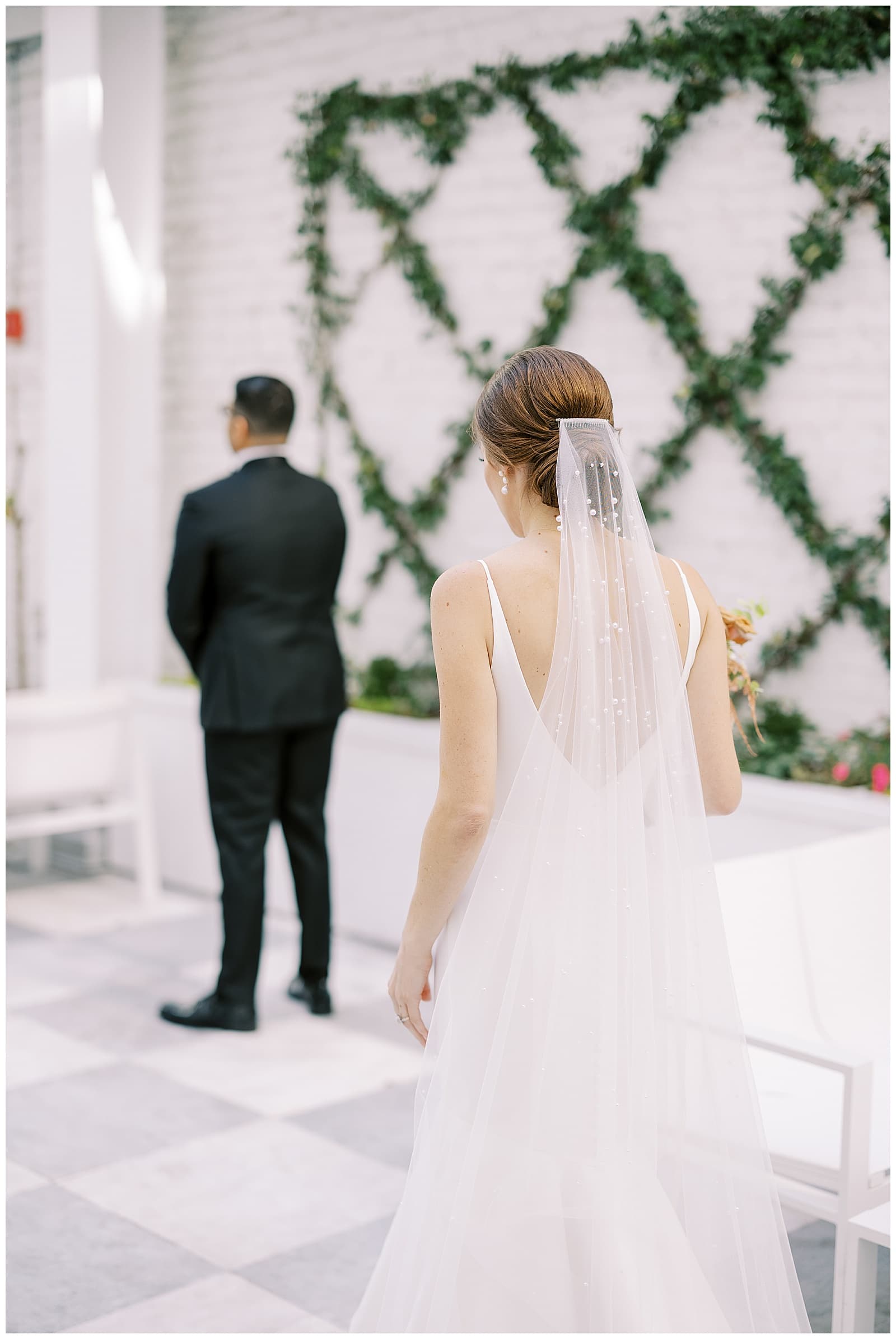 The back view of a bride in a white wedding dress with a long white veil walking across the courtyard of the Quirk hotel richmond to a young man in a black tuxedo standing away from her 