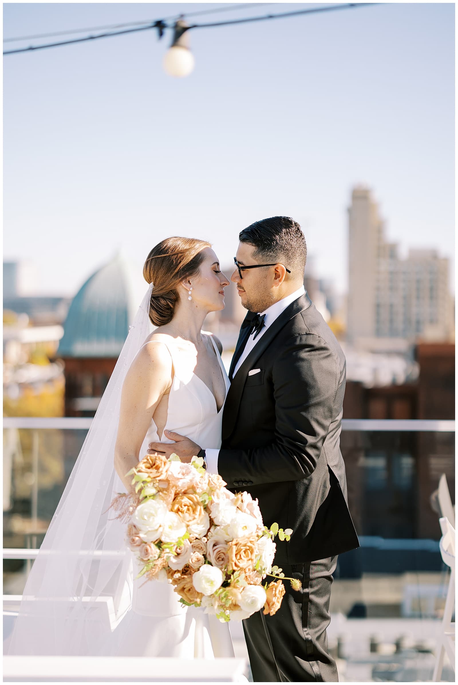 A red headed woman in a white modern wedding dress with a veil leans in to kiss a handsome groom in a black tuxedo with the Richmond skyline behind them on the roof of the Quirk Hotel Richmond