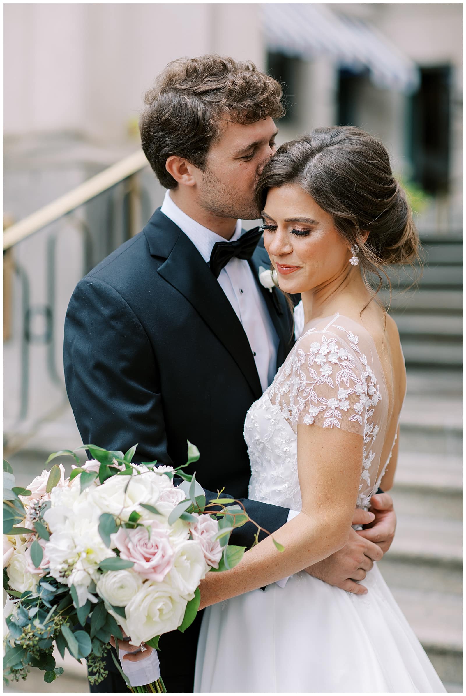 A young man with brown hair in a black bow tie and tuxedo is embracing a young brown haired woman in a white wedding gown on the steps of the Willard Hotel DC