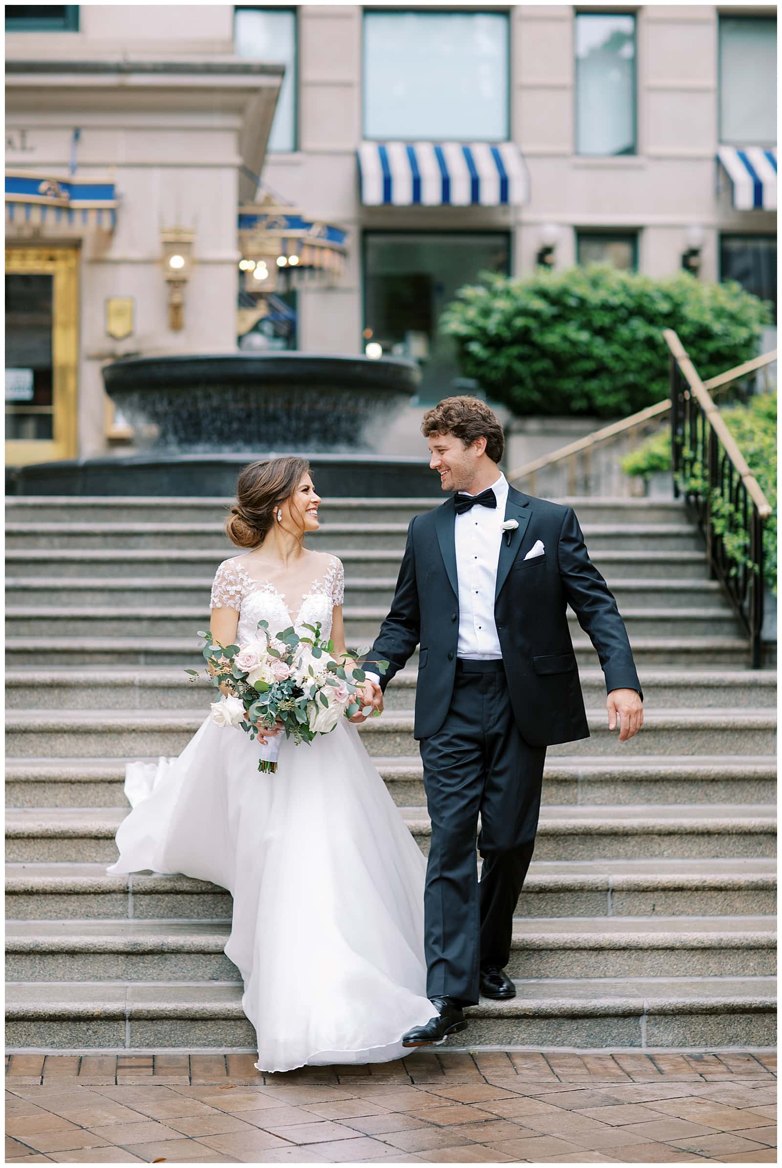 A Young Brown haired woman in a white wedding dress holding a large floral bouquet holds the hand of a young man in a black tuxedo smiling as they walk down the steps outside of the Willard Hotel DC