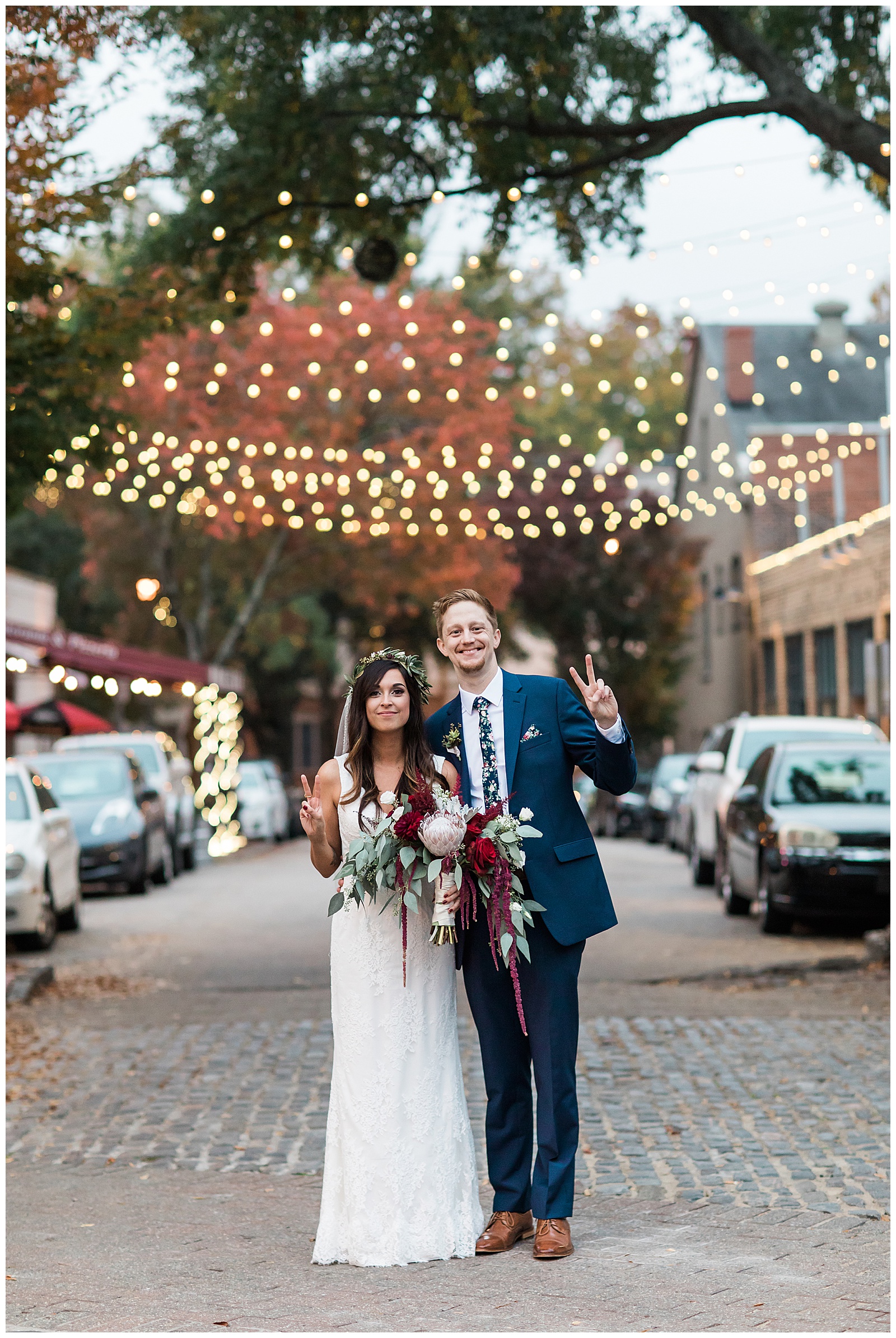A dark haired woman in a white wedding gown stands beside a tall red headed gentleman in a blue suit outside of Market hall Raleigh while holding up the peace sign with their fingers