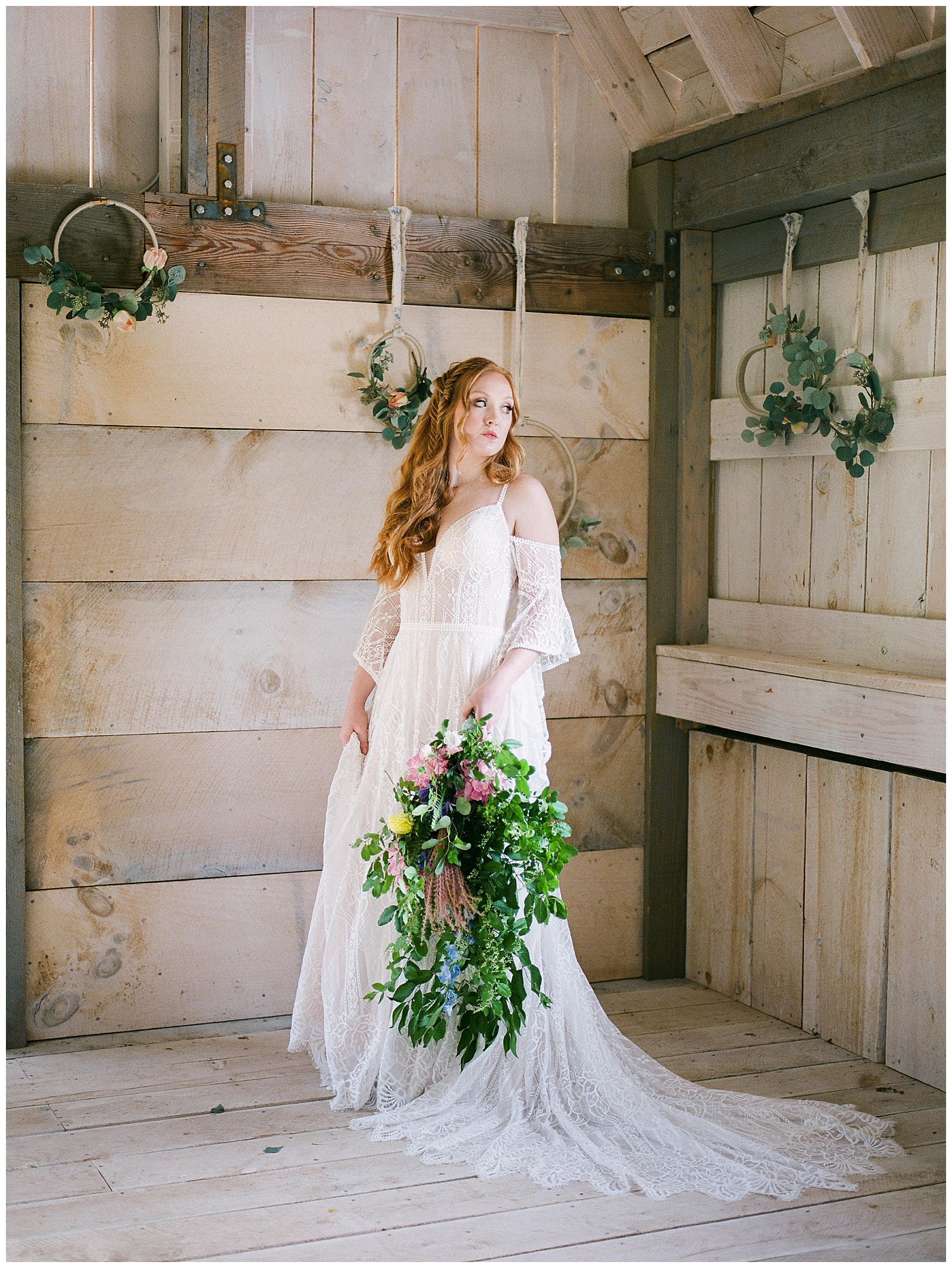 Redheaded woman in a bohemian white bridal gown stands in Ramble Creek Vineyard barn holding a large floral bouquet looking out the window