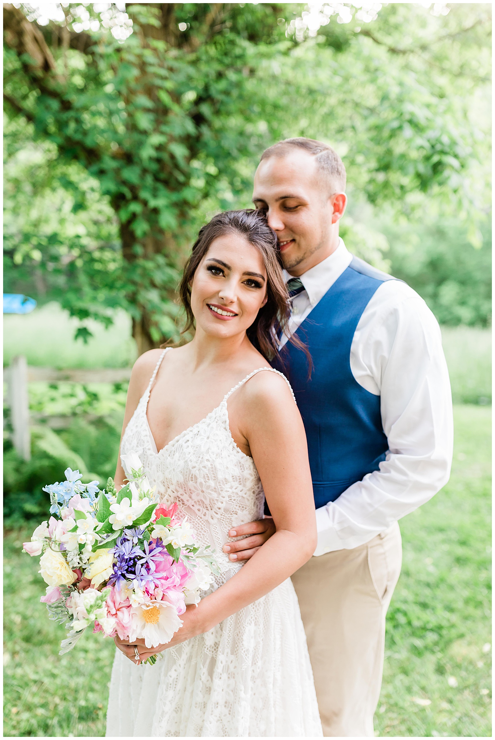 A dark haired woman smiles directly at the camera in a white wedding dress with a large bouquet of flowers while a tall man in a suit stands behind her embracing her and kissing her temple outside at The Side Porch in Johnson City