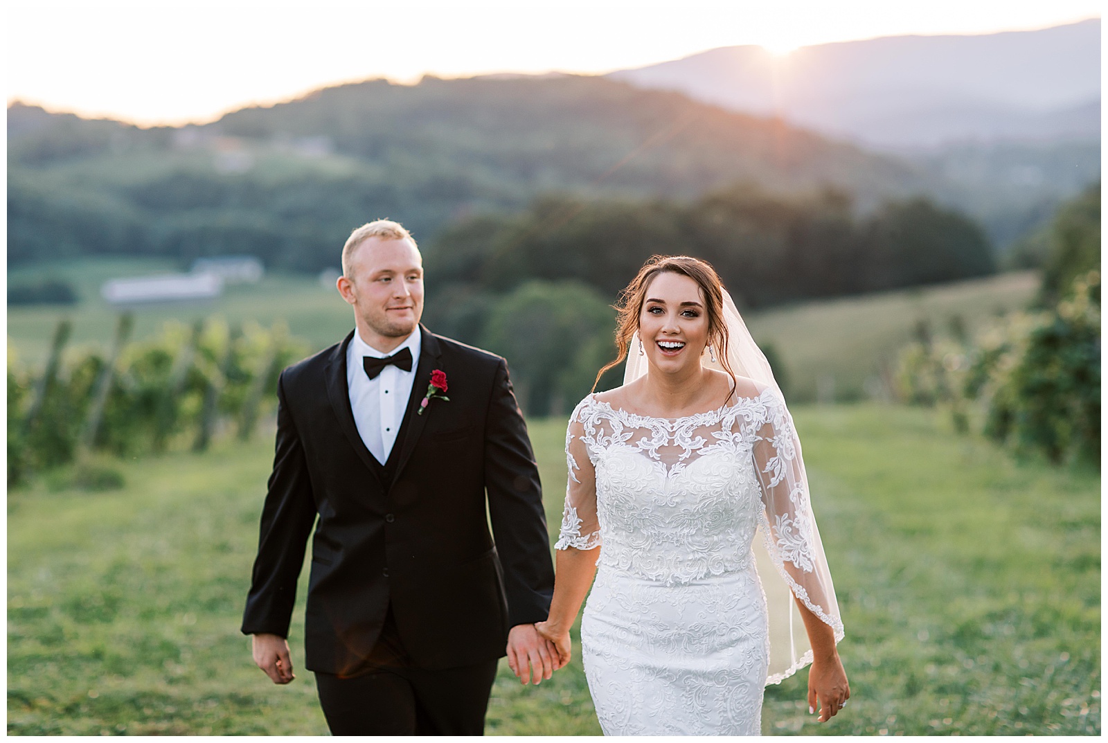 A young woman with dark brown hair laughs while holding a tall blonde man in a black tux's hand while they stroll through the vineyard at sunset