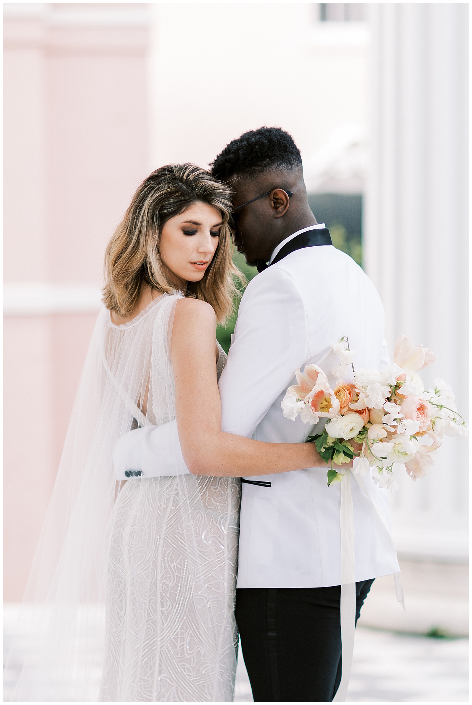 Blonde haired woman with a white wedding dress and tulle cape is embraced by a dark skinned man in a white tuxedo on the porch of the william aiken house in charleston sc