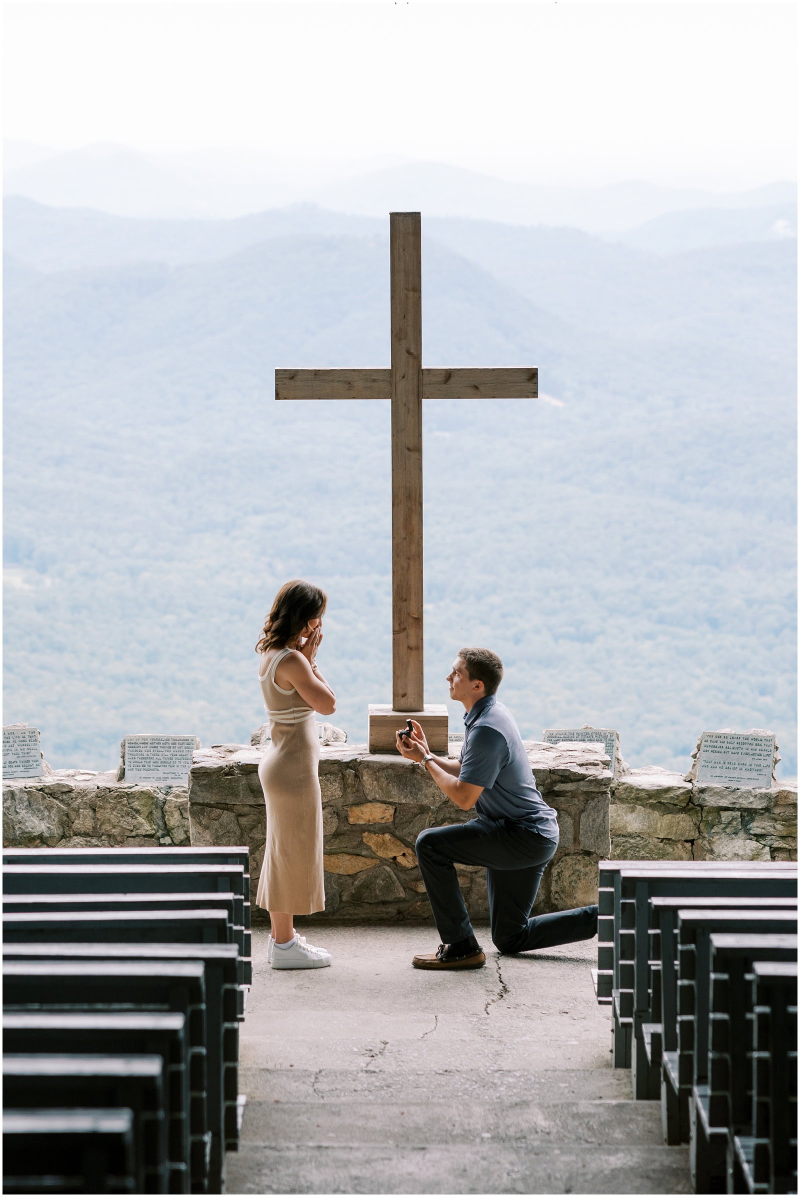 Beautiful mountain wedding proposal with a man kneeling offering a ring in front of a large wooden cross at an outdoor scenic overlook. Romantic moment with stunning mountain views in the background.