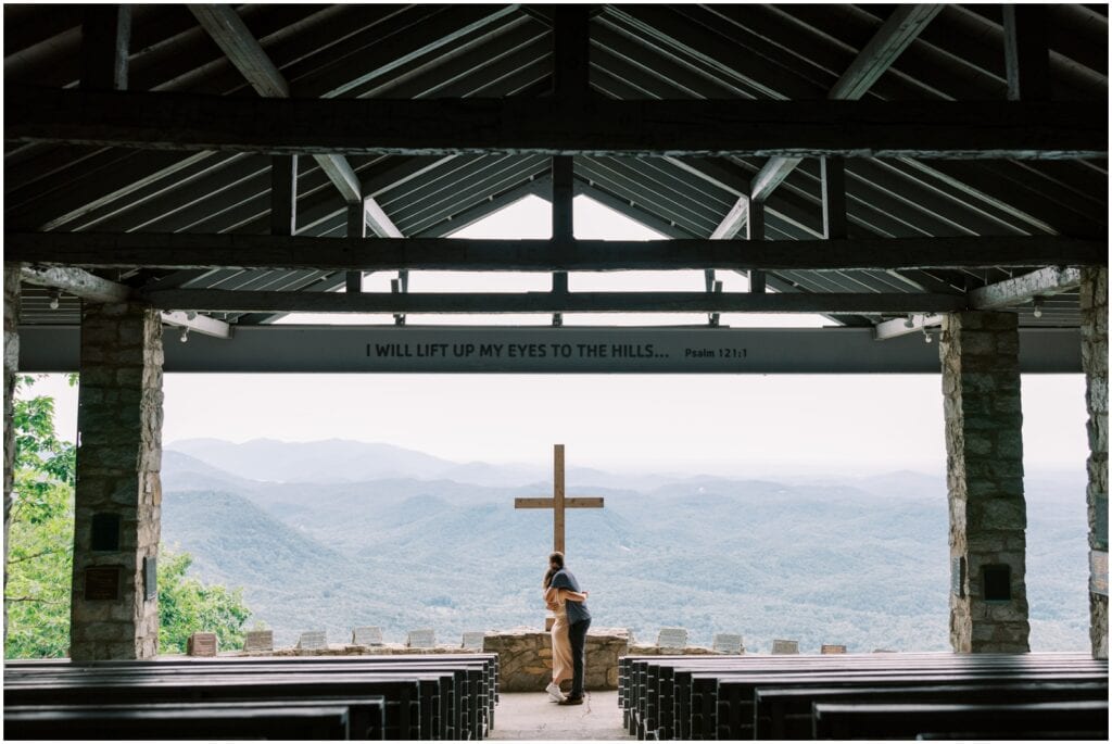 Serene church interior with a view of distant mountains, featuring a wooden cross and a couple embracing, with overhead beams and natural light, symbolizing faith and devotion.